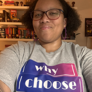 Photo of a Black woman wearing a gray tee shirt with the Why Choose: A Romance Bookstore purple and pink logo